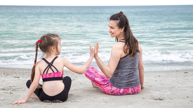 Una joven madre con una pequeña hija en ropa deportiva está sentada en la playa con el fondo del mar. valores familiares y estilo de vida saludable.