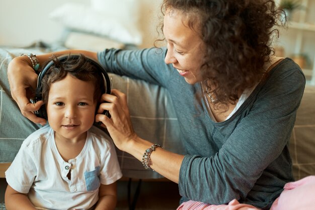 Joven madre de raza mixta cuidando a su adorable hijo de tres años, usando auriculares inalámbricos