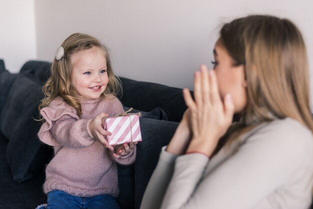 La joven madre sentada en el sofá en casa recibió una sorpresa de su hija mirando el regalo con asombro en el día de la madre