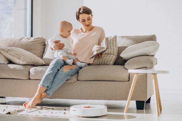 Foto gratuita joven madre sentada en un sofá con un hijo pequeño y viendo robot aspirador haciendo las tareas del hogar