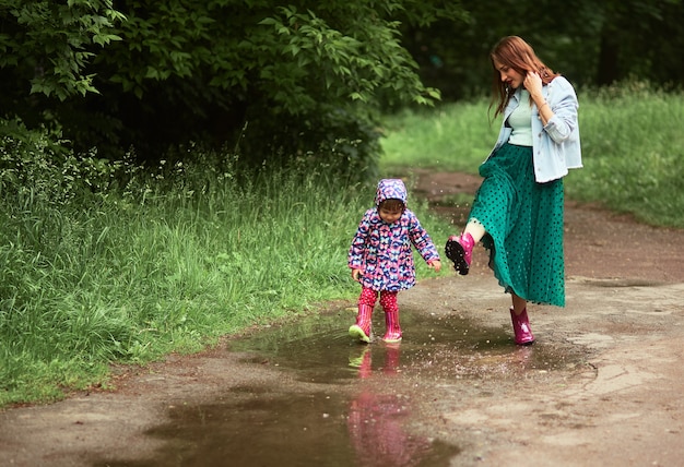 Joven madre y su pequeña hija se divierten caminando en botas de goma en las piscinas en el parque