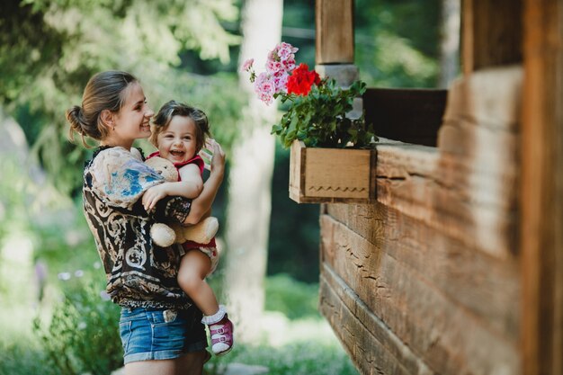 Joven madre tiene niño pequeño alegre de pie con ella antes de casa de madera vieja