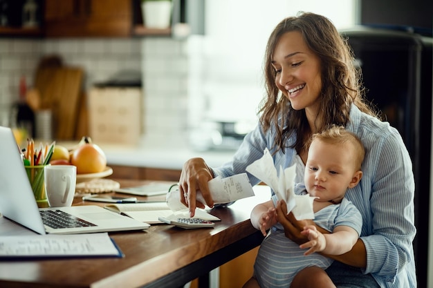 Foto gratuita joven madre trabajadora con bebé usando calculadora y revisando facturas mientras calcula las finanzas del hogar