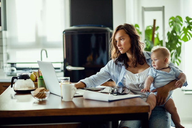 Foto gratuita joven madre trabajadora que usa la computadora mientras sostiene a su bebé y planifica las finanzas del hogar