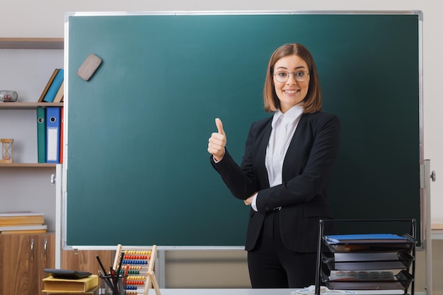 Joven maestra con anteojos parada cerca de la pizarra en el aula explicando la lección mostrando el pulgar hacia arriba sonriendo alegremente feliz y positiva