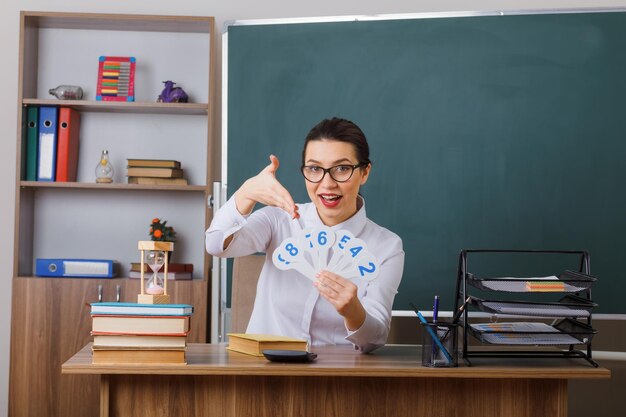 Joven maestra con anteojos presentando matrículas explicando la lección feliz y positiva sonriendo sentada en el escritorio de la escuela frente a la pizarra en el aula