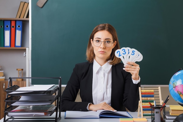 Joven maestra con anteojos sentada en el escritorio de la escuela frente a la pizarra en el aula sosteniendo placas de matrícula explicando la lección con cara seria