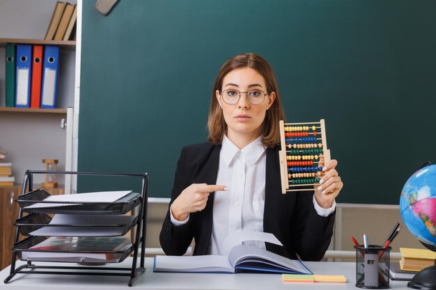 Joven maestra con anteojos sentada en el escritorio de la escuela frente a la pizarra en el aula usando ábaco apuntando con el dedo índice con confianza