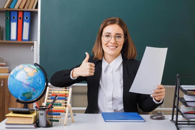 Joven maestra con anteojos sentada en el escritorio de la escuela con globo y libros frente a la pizarra en el aula sosteniendo una hoja de papel blanca vacía mostrando el pulgar hacia arriba sonriendo alegremente