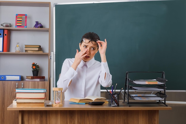 Foto gratuita joven maestra con anteojos sentada en el escritorio de la escuela con un libro frente a la pizarra en el aula con aspecto cansado y con exceso de trabajo