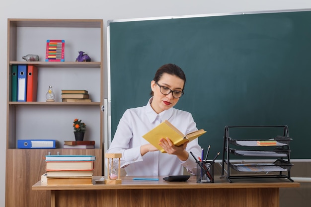 Joven maestra con anteojos sentada en el escritorio de la escuela con un libro frente a la pizarra en el aula explicando la lección con confianza