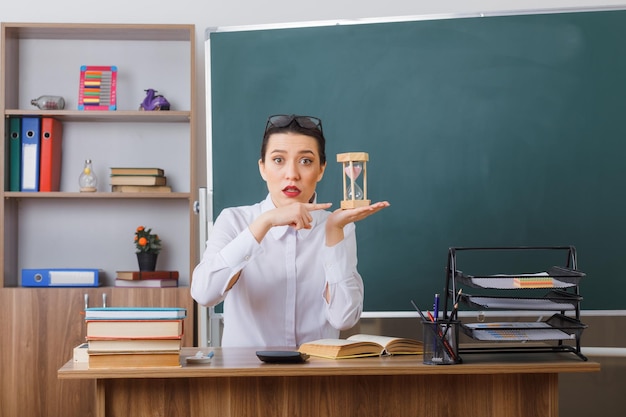 Foto gratuita joven maestra con anteojos sentada en el escritorio de la escuela con un libro frente a la pizarra en el aula presentando un reloj de arena sorprendido