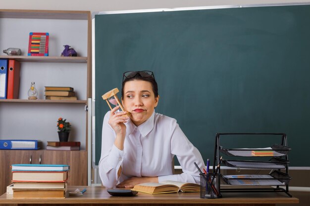 Joven maestra con anteojos sentada en el escritorio de la escuela con un libro frente a la pizarra en el aula sosteniendo un reloj de arena con aspecto cansado y aburrido