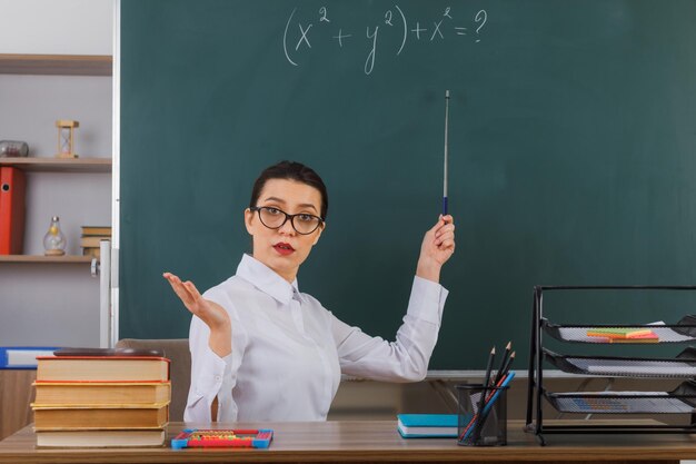 Joven maestra con anteojos sosteniendo un puntero mientras explica la lección con confianza sentada en el escritorio de la escuela frente a la pizarra en el aula