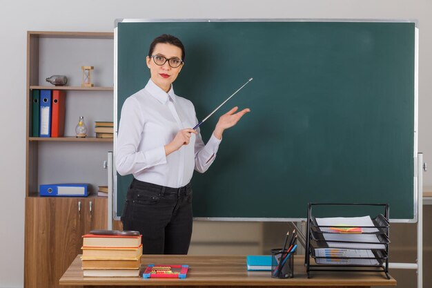 Una joven maestra con anteojos sosteniendo un puntero mientras explica la lección luciendo confiada de pie en el escritorio de la escuela frente a la pizarra en el aula