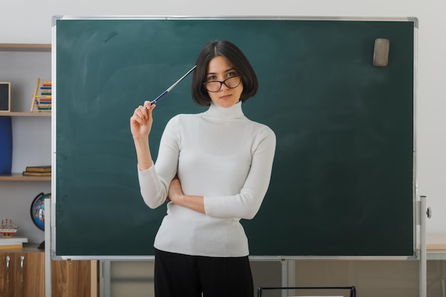 joven maestra complacida con anteojos parada frente a la pizarra sosteniendo el puntero en la cabeza en el aula