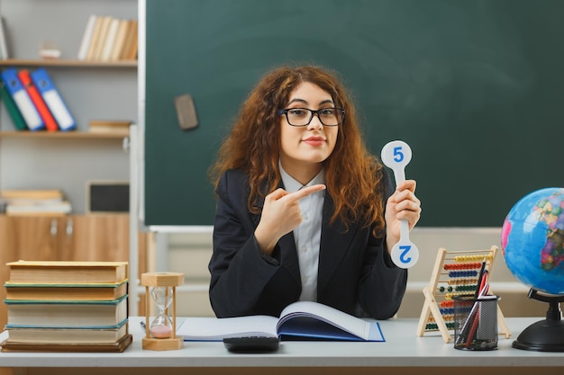 joven maestra complacida con gafas sosteniendo y señalando el ventilador sentado en el escritorio con herramientas escolares en el aula