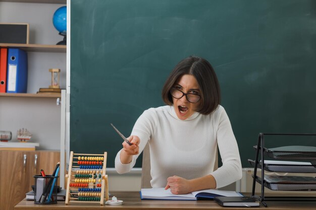una joven maestra enojada que usa anteojos apunta a la cámara con un puntero sentado en el escritorio con herramientas escolares en el aula