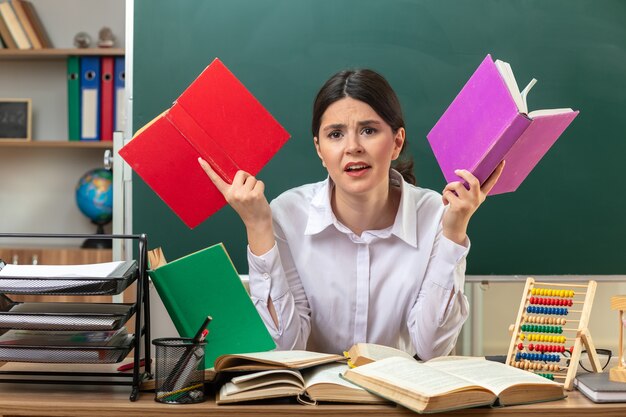 Joven maestra insatisfecha sosteniendo libro sentado a la mesa con herramientas escolares en el aula