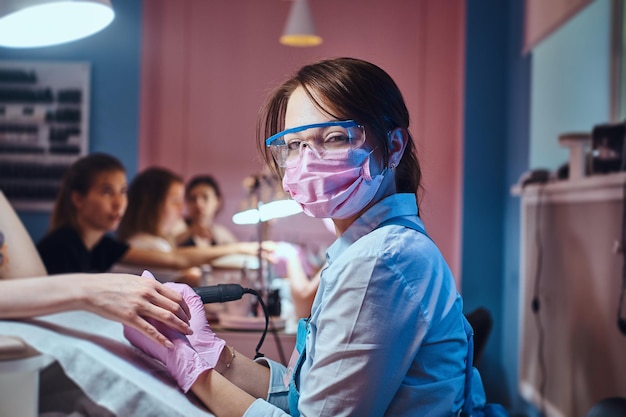 Joven maestra de manicura alegre en su propio lugar de trabajo con el cliente está trabajando en las uñas de la mujer.