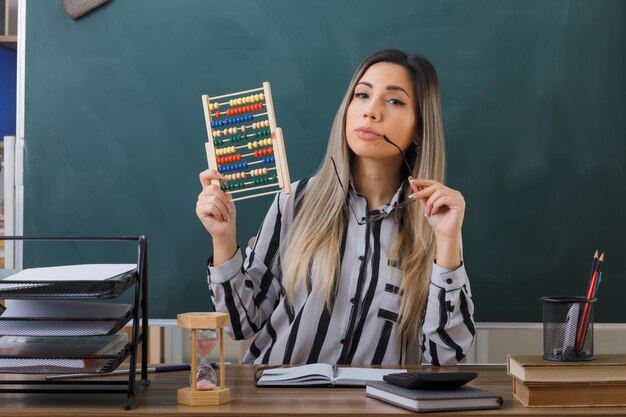 joven maestra sentada en el escritorio de la escuela frente a la pizarra en el aula explicando la lección sosteniendo abacus mirando a la cámara intrigada