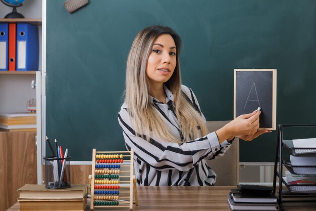 joven maestra sentada en el escritorio de la escuela frente a la pizarra en el aula explicando la lección sosteniendo una pequeña pizarra mirando a la cámara sonriendo confiada