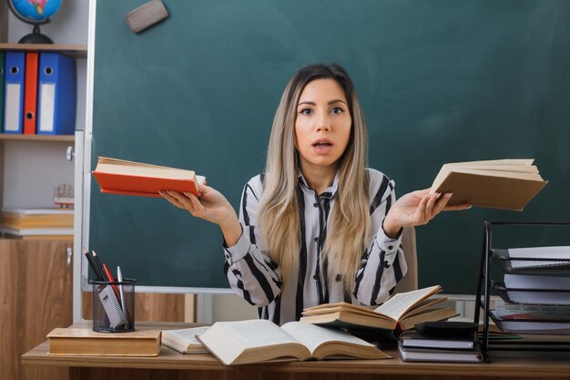 joven maestra sentada en el escritorio de la escuela frente a la pizarra en el aula sosteniendo libros mirando a la cámara confundida tratando de hacer una elección