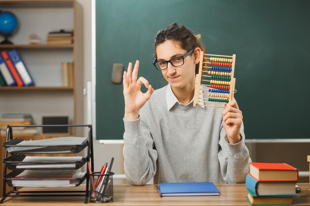 un joven maestro confiado apunta a la pizarra sosteniendo un ábaco mostrando un gesto correcto sentado en el escritorio con herramientas escolares en el aula