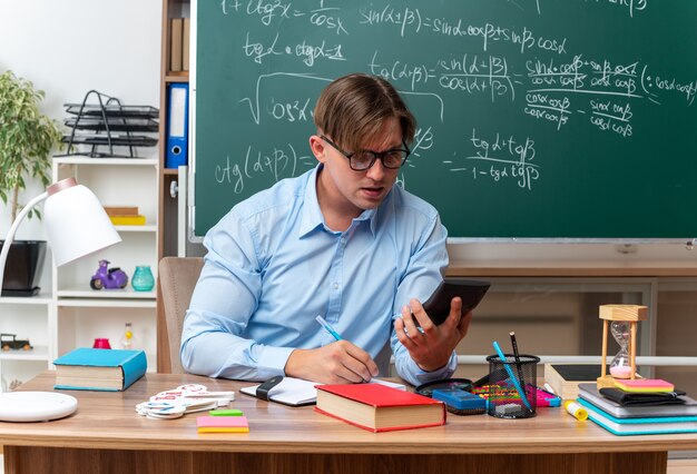 Joven maestro con gafas mirando su teléfono inteligente mirando confiado sentado en el escritorio de la escuela con libros y notas frente a la pizarra en el aula