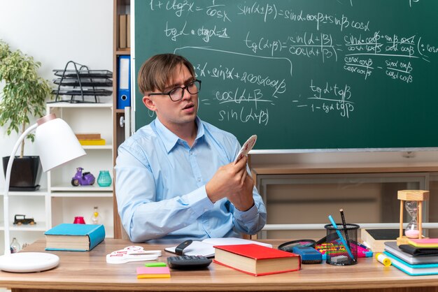 Joven maestro con gafas preparando la lección mirando confiado sentado en el escritorio de la escuela con libros y notas frente a la pizarra en el aula