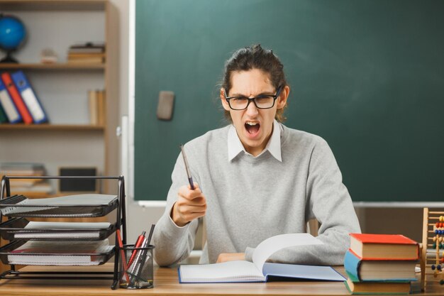 joven maestro con gafas sosteniendo un puntero sentado en el escritorio con herramientas escolares en el aula