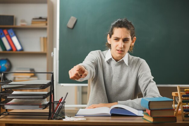 un joven maestro pensante apunta a la cámara sentado en el escritorio con herramientas escolares en el aula