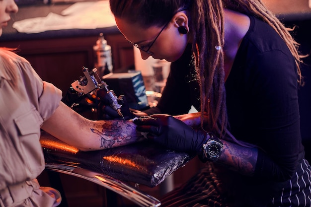 El joven maestro de tatuajes enfocado está haciendo tatuajes en la mano del cliente en el salón de tatuajes oscuros.