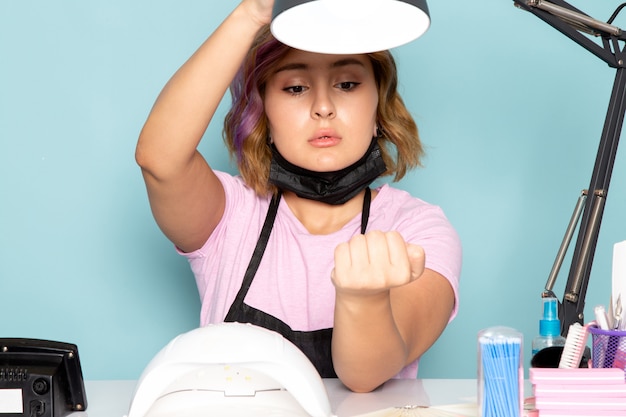 Una joven manicura de vista frontal en camiseta rosa con guantes negros y máscara negra sentada frente a la mesa revisando sus uñas en azul