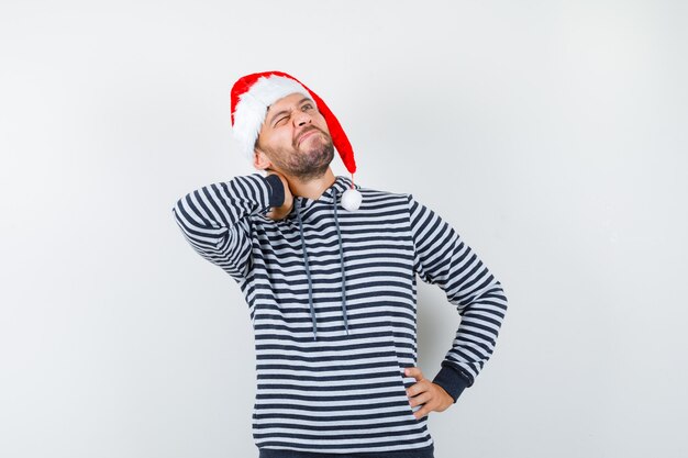 Joven manteniendo la mano en el cuello, mirando hacia arriba con capucha, gorro de Papá Noel y mirando pensativo,