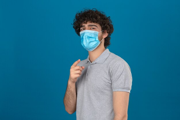 Joven en máscara protectora médica apuntando a la cámara con mirada de confianza sobre fondo azul aislado