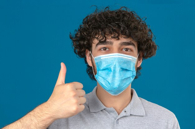 Joven en máscara protectora médica mirando a la cámara con cara feliz mostrando el pulgar hacia arriba sobre fondo azul aislado