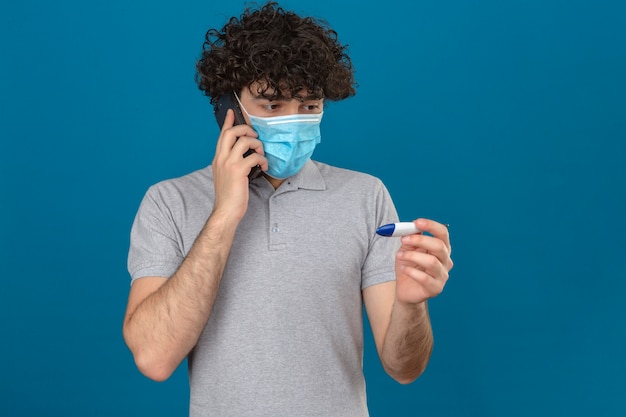 Foto gratuita joven en máscara protectora médica mirando el termómetro digital en la mano mirando nervioso sobre fondo azul aislado