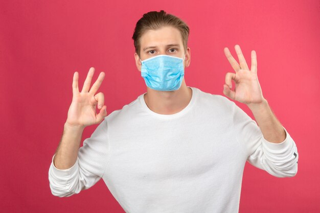 Joven en máscara protectora médica mostrando signos de ok con los dedos y las manos mirando a la cámara sobre fondo rosa aislado