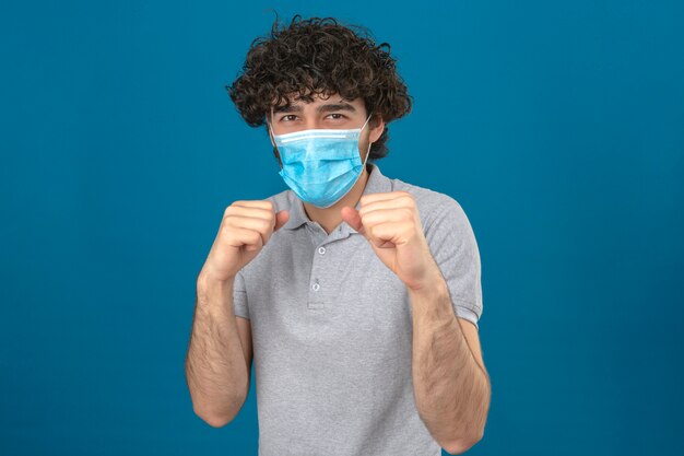 Joven en máscara protectora médica de pie en pose de boxeador con puño listo para luchar sobre fondo azul aislado