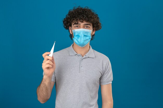 Joven en máscara protectora médica sosteniendo un termómetro digital en la mano mirando a la cámara con cara seria sobre fondo azul aislado
