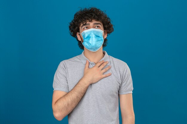 Joven en máscara protectora médica tocando el pecho para comprobar el pulmón mientras respira sobre fondo azul aislado