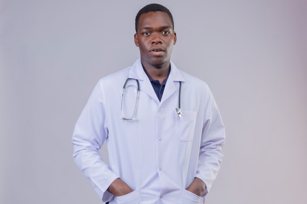 Joven médico afroamericano vistiendo bata blanca con estetoscopio con las manos en los bolsillos con expresión seria de confianza