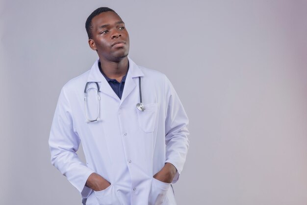 Joven médico afroamericano vistiendo bata blanca con estetoscopio con las manos en los bolsillos mirando a un lado con expresión seria y segura en la cara