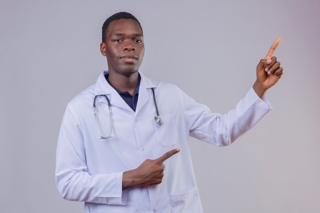 Joven médico afroamericano vistiendo bata blanca con estetoscopio mirando confiado apuntando con los dedos índices de ambas manos hacia el lado