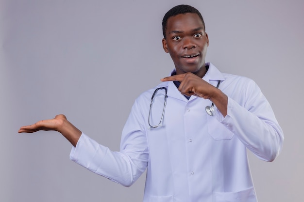 Foto gratuita joven médico afroamericano vistiendo bata blanca con estetoscopio mirando sorprendido y feliz apuntando con el dedo hacia el lado que presenta con el brazo oh su mano