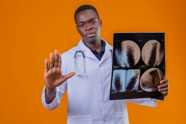 Joven médico afroamericano vistiendo bata blanca con estetoscopio sosteniendo radiografía de los pulmones haciendo señal de pare con la mano abierta con cara seria