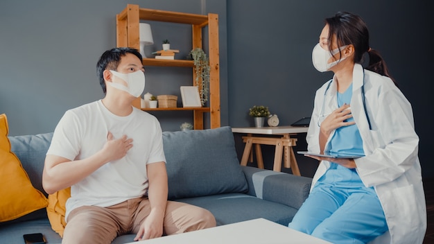 Joven médico de Asia mujer médico usa mascarilla usando tableta digital compartiendo buenas noticias sobre pruebas de salud con paciente masculino feliz sentado en el sofá en casa.