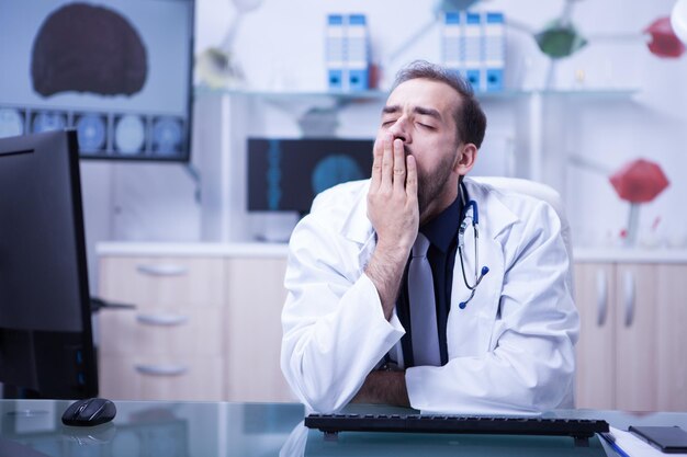 Joven médico barbudo cansado y bostezando sentado en la oficina en su gabinete de hospital. Médico estresado.