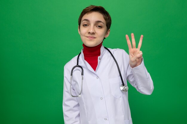 Joven médico en bata blanca con estetoscopio alrededor del cuello mirando al frente feliz y positivo sonriendo confiado mostrando el número tres con los dedos de pie sobre la pared verde